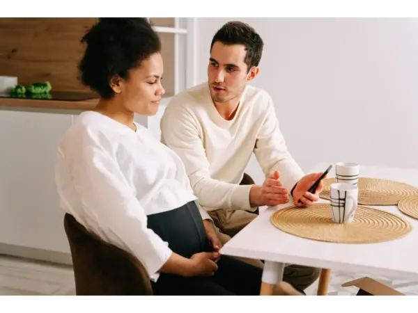 An interracial couple discussing having kids and pregnancy plans while sitting together at a kitchen table and sharing a thoughtful conversation.