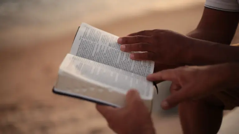 In Wednesday's daily devotional, two men's hands are seen holding a Bible, seemingly discussing it, near the ocean.