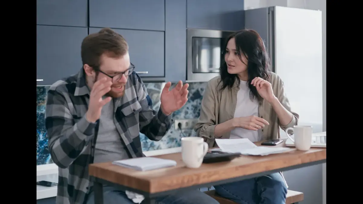 A man and a woman sitting at a kitchen counter engaged in an animated conversation, with papers and coffee mugs in front of them, Couple arguing about money, budgeting, finances