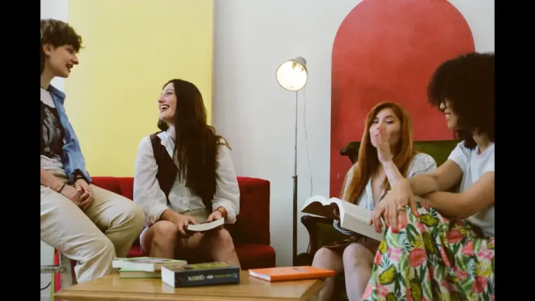 A group of four friends sitting together on a couch, smiling and laughing while holding books in a cozy, colorful room. book club, books, reading