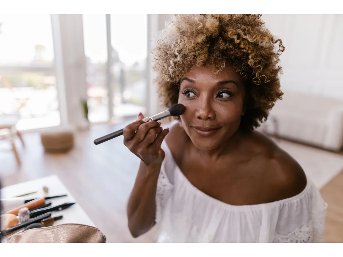 Woman with curly hair applying cherry cola makeup in a well-lit room, creating a natural yet polished beauty look.