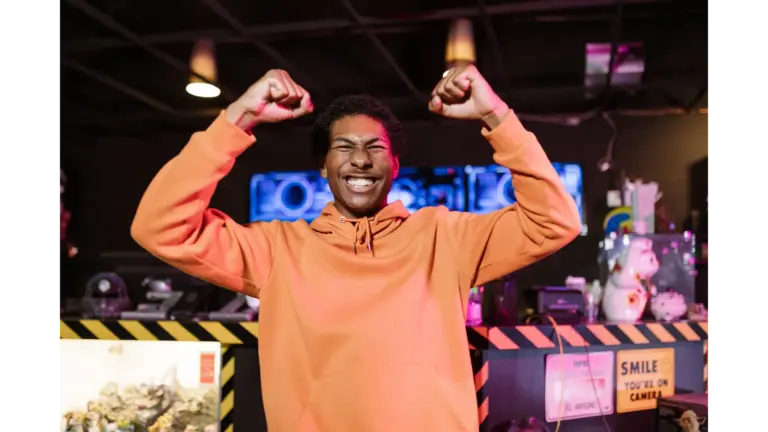 A cheerful young man in an orange hoodie celebrates a gaming victory indoors with a big smile, reflecting the excitement often seen before major game show scandals change television history.