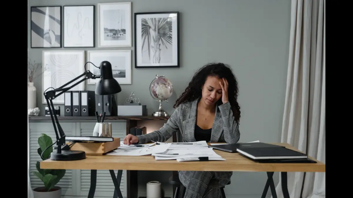 A woman sitting at a desk in a home office, looking stressed while holding her forehead, surrounded by paperwork and a desk lamp. How to make decisions when you feel overwhelmed. Decision making when overwhelmed.