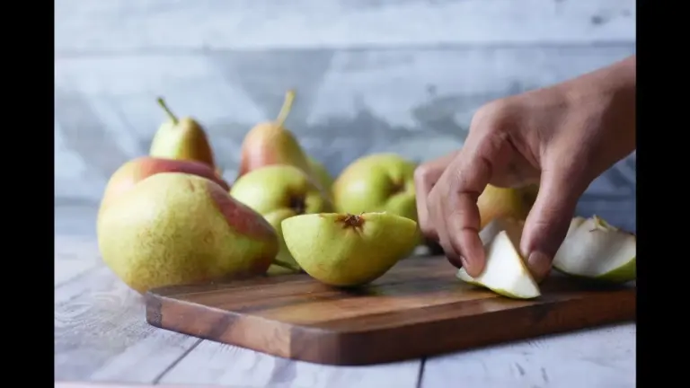 A hand slices a pear on a wooden cutting board, surrounded by whole pears in the background.