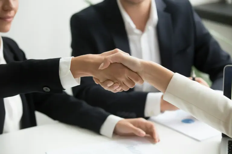 Two individuals shaking hands in a business meeting, with another person in a suit visible in the background. Papers and a chart are on the table.