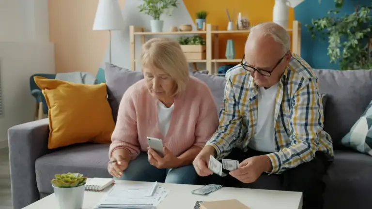 A senior couple is sitting on a couch, discussing financial documents while one of them holds a phone and the other is examining a receipt. retirement savings, early retirement, FIRE movement